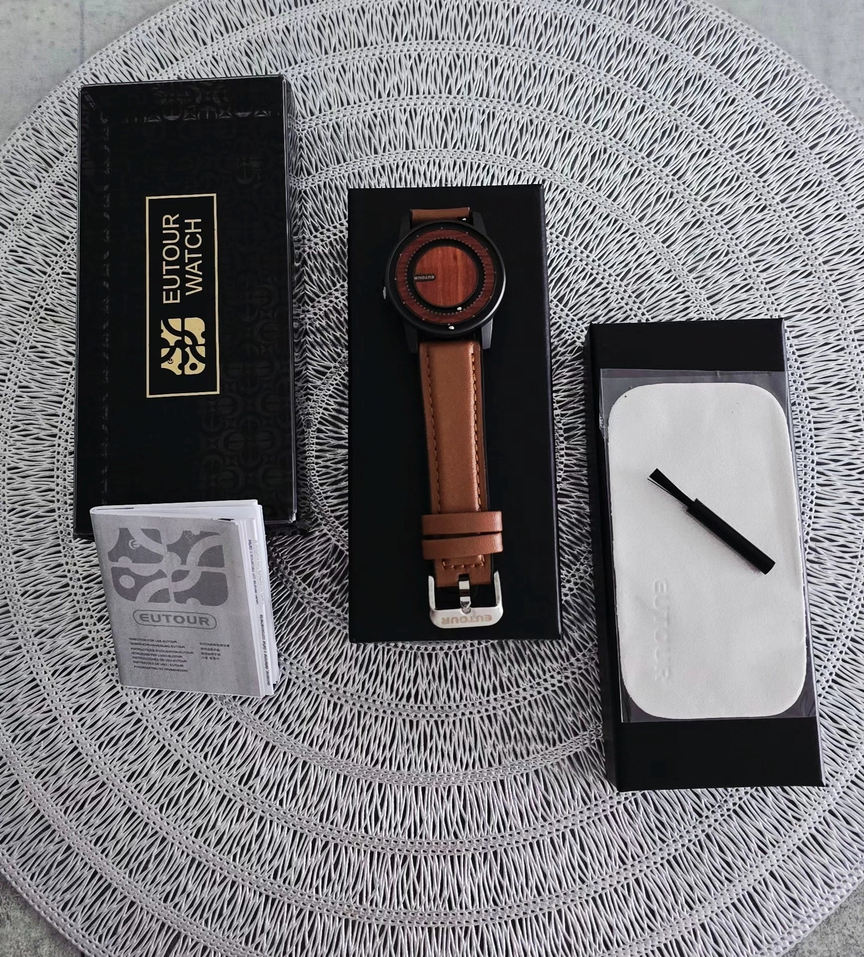 A wristwatch with a brown leather strap and round black case displayed on a black presentation box, alongside its black branded box, a small booklet, and a white cleaning cloth on a woven round mat.