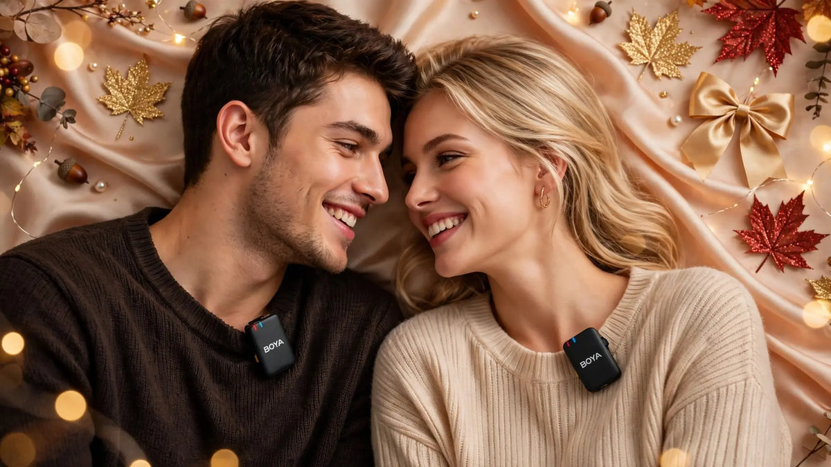 Another young couple young man with light brown hair and beard in chocolate brown sweater and beautiful young girl with long dark wavy hair in beige sweater smiling at each other, only small exact black BOYA BOYALINK wireless transmitter mics clipped to sweaters no case, background champagne silk draped gold glitter maple leaves red glitter maple acorns pearls gold satin bows fairy lights bokeh autumn luxury lifestyle banner no text - Goldener Wahl