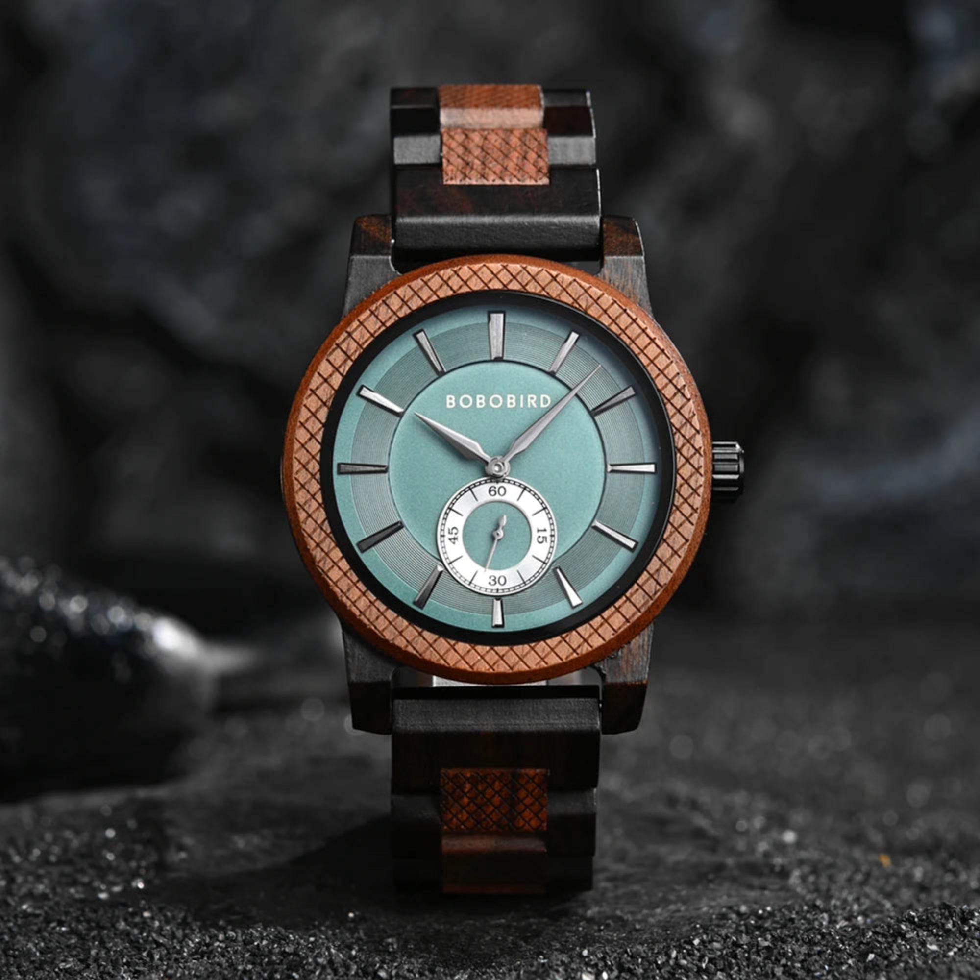 A wooden watch with a green dial and brown bands, displayed against a dark textured background with stones and a branch.