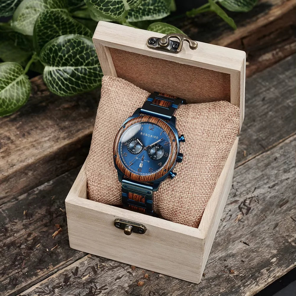 A wristwatch with a blue dial and blue metal bracelet displayed inside an open light wood presentation box with a burlap cushion, set on a rustic wooden surface with green leaves in the background.