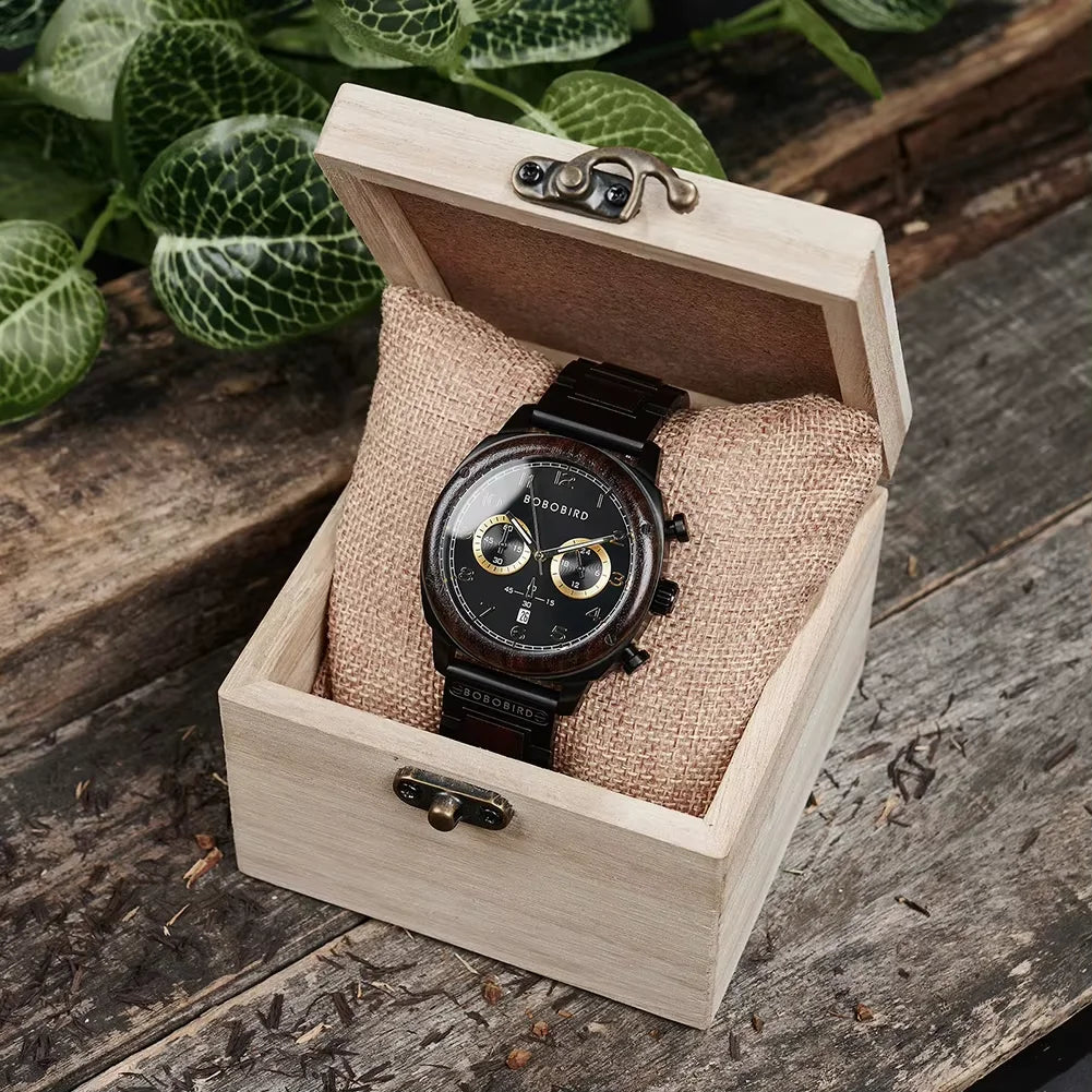 A black analog watch with a wood-grain dial and black strap, presented inside an open light wood gift box with a burlap insert, set on a rustic wooden surface with green leaves in the background.