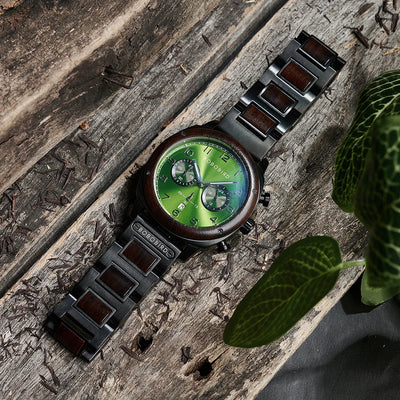 A wristwatch with a green dial and dark brown wood-tone accents on a weathered wooden surface, styled with a green leaf in the foreground.