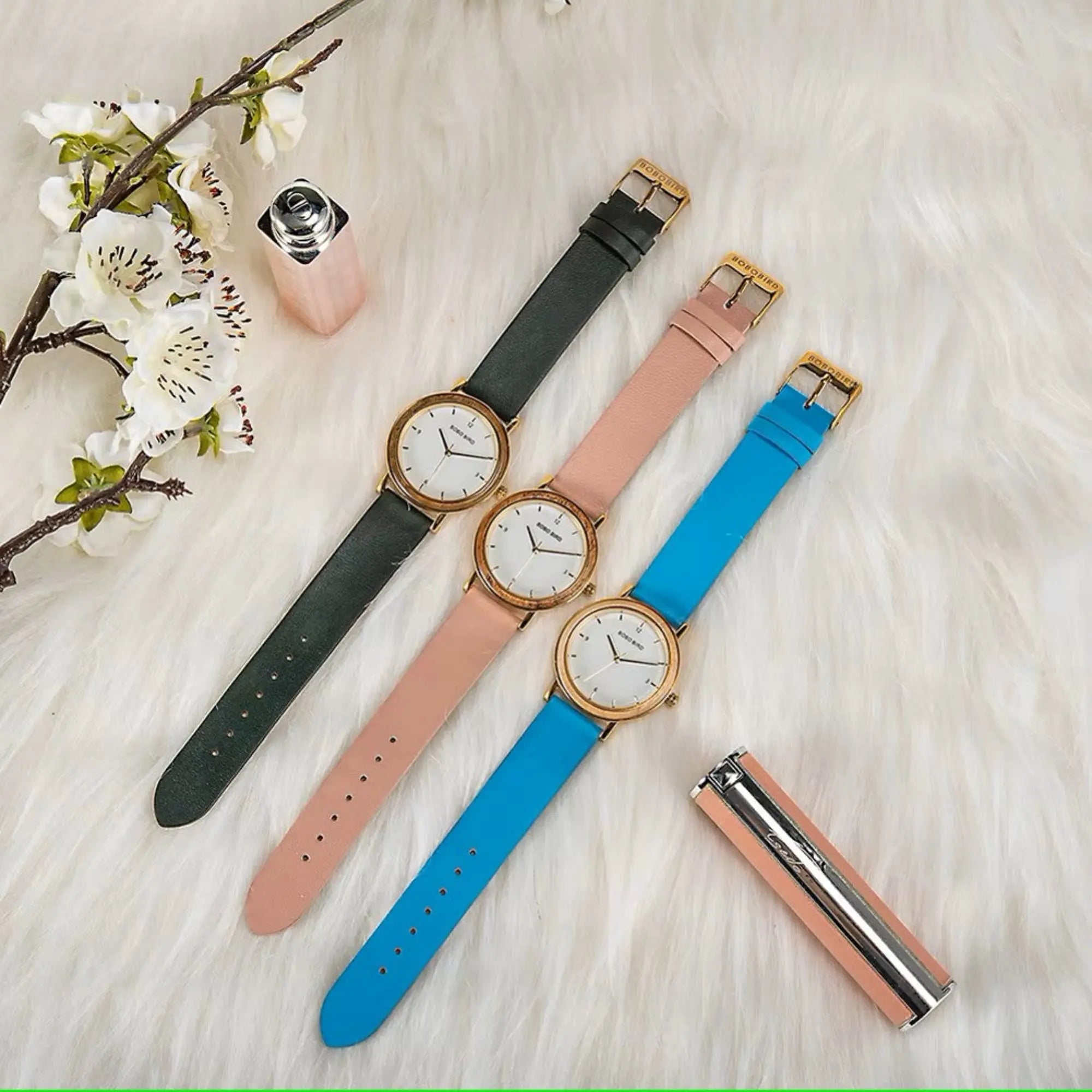 Three watches with different colored straps on a light surface with flowers and a bottle.