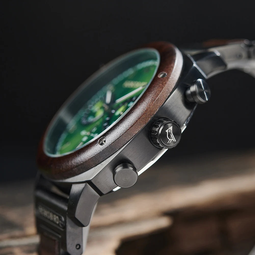 A close-up side view of a rugged chronograph watch with a dark wood bezel, matte black metal case, and a green dial, resting on a rustic wooden surface against a dark background.