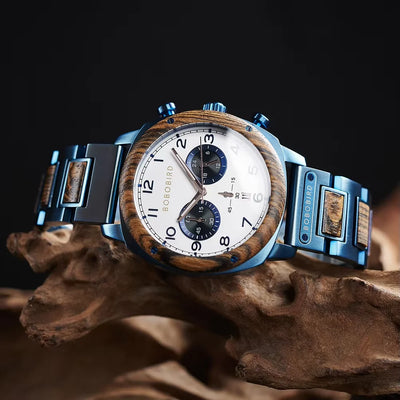 Boboiprin men's watch with a blue stainless steel case and link bracelet featuring wood inlays, a white dial with black numerals, and two blue subdials, displayed on a piece of driftwood against a dark background.
