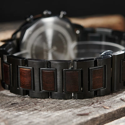 Close-up of a watch bracelet with dark gunmetal links and rectangular brown wood inlays, resting on a rustic wooden surface with the watch case blurred in the background.