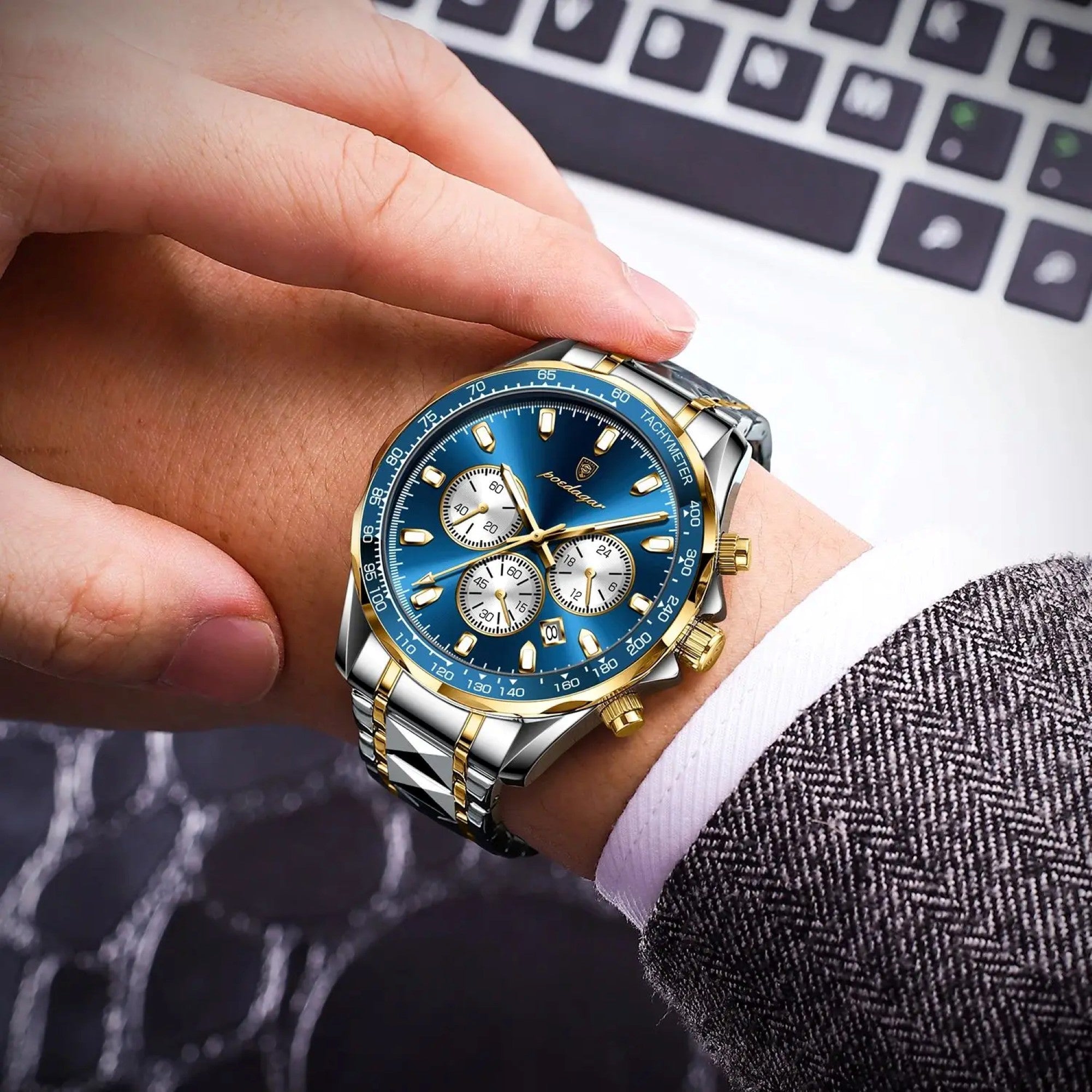 Hand wearing a blue and gold watch with a blurred laptop background