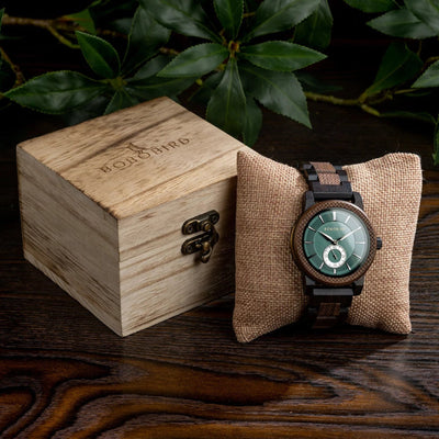 Wooden watch with green face in a wooden box and on a textured pillow with green leaves in the background.