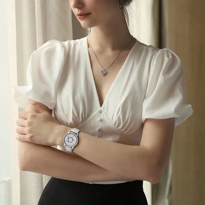 Woman wearing a white blouse with puffed sleeves, black pants, and a silver watch, standing against a neutral background.