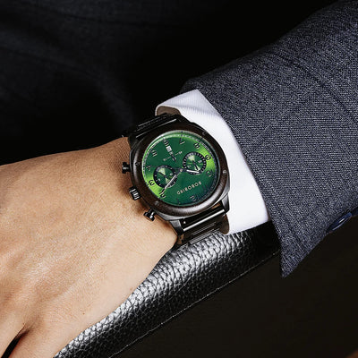 A close-up of a wrist wearing a SONDOR chronograph watch with a green dial and black case, shown on a person's wrist with a dark gray suit sleeve and black textured surface in the background.