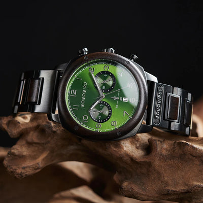 A wristwatch with a green dial, black case, and black metal bracelet, resting on a piece of natural wood against a dark background. The brand name BOBOIRD is visible on the watch dial and bracelet.