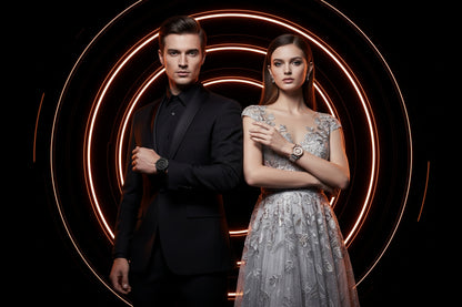 Man in black suit and woman in silver dress standing against a dark background with circular light patterns.