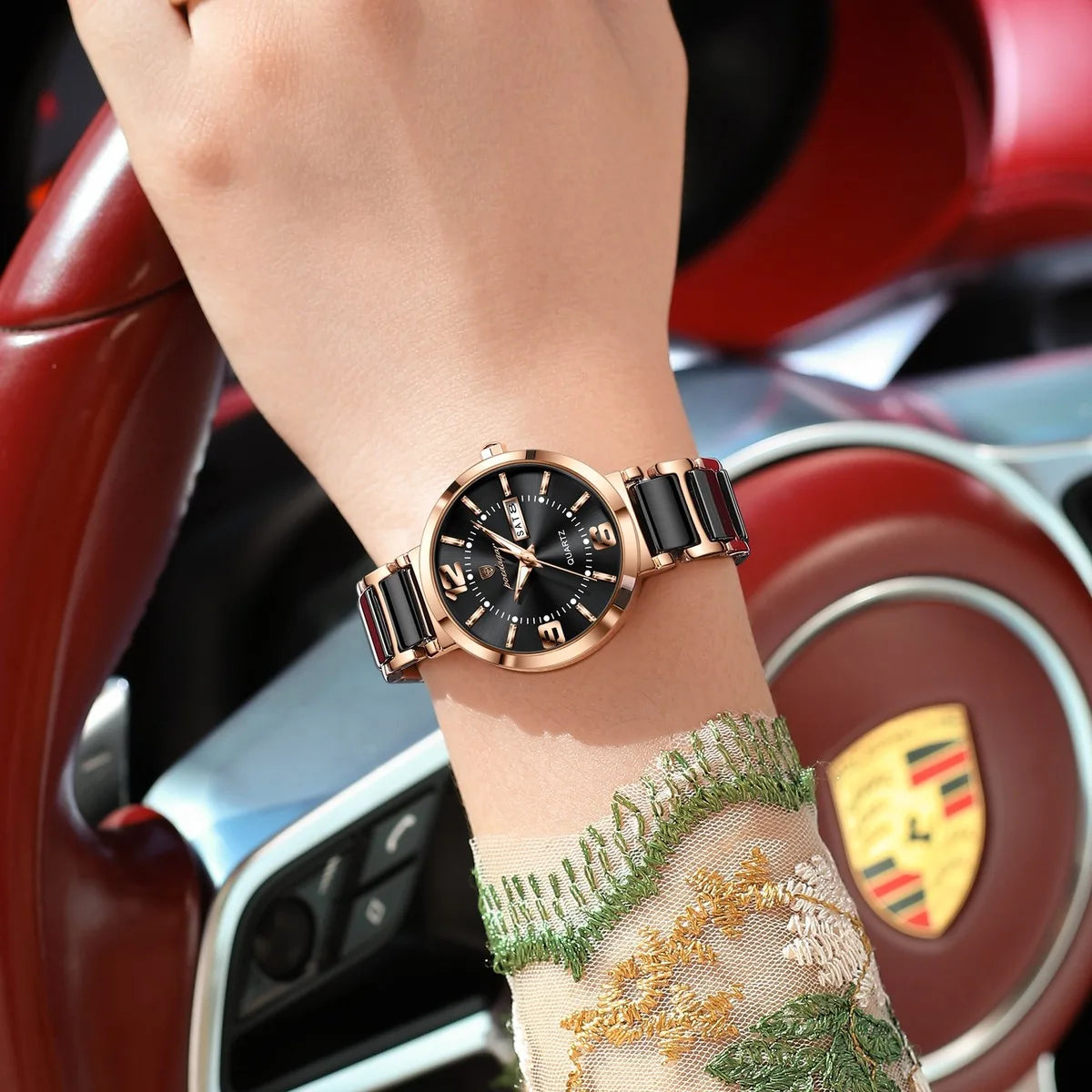 A close-up of a woman's wrist wearing a Porsche-branded watch with a black dial and a black-and-gold ceramic link band, posed in front of a red car interior with the Porsche logo visible.