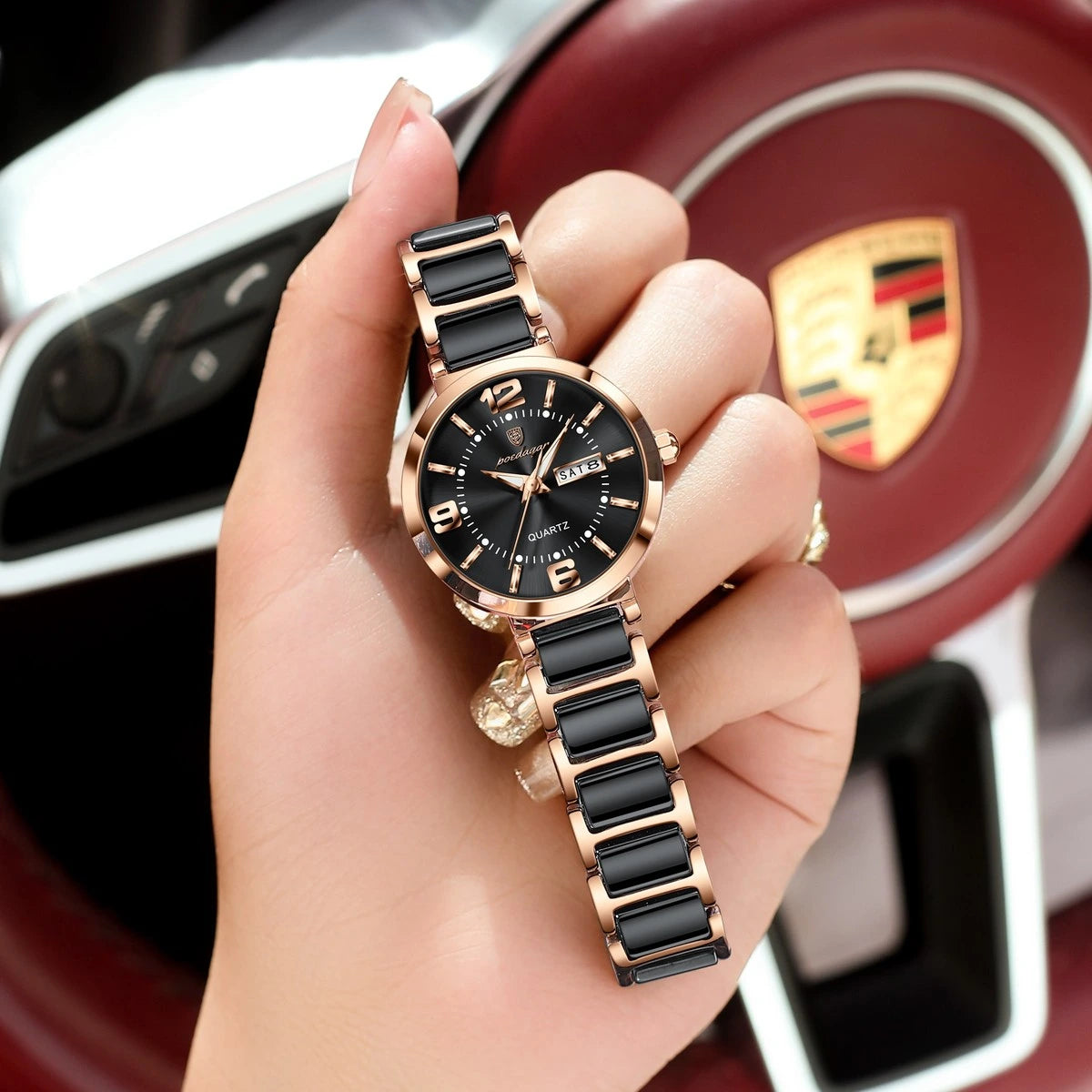 A close-up of a hand holding a wristwatch with a black link strap and a black round dial with rose gold-toned case details, set against a blurred Porsche steering wheel background.